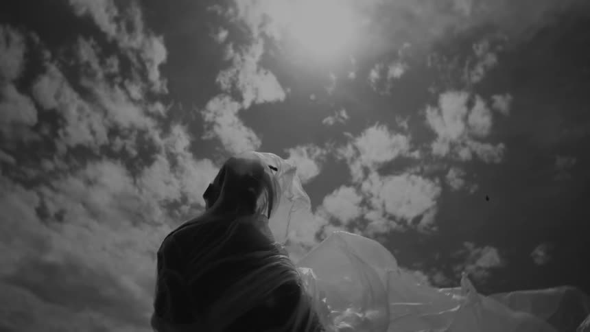 A person is wrapped in plastic with the plastic billowing out behind them set against a cloudy sky