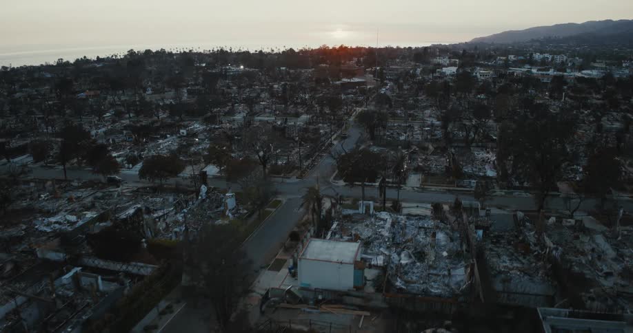 A neighborhood is devastated by fire with charred remains of buildings and trees under a setting sun