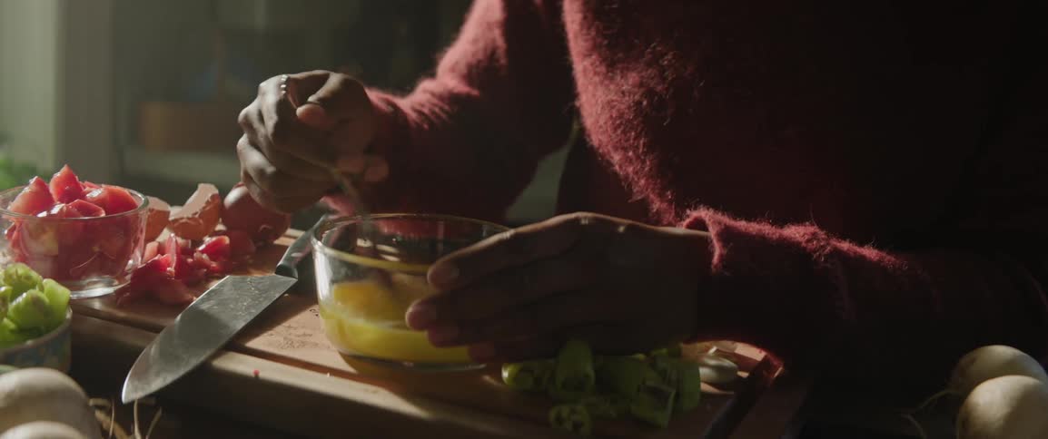 A person wearing a red sweater is whisking eggs in a glass bowl on a wooden cutting board surrounded by chopped green vegetables and a knife