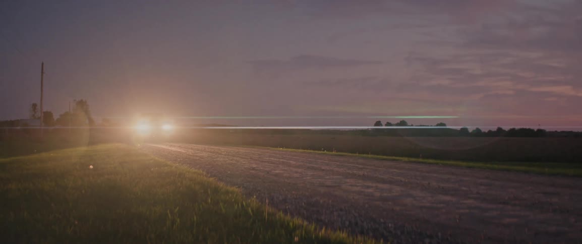 A car with its headlights on is driving along a rural dirt road at dusk with a field and a line of trees visible in the background under a purple and pink sky