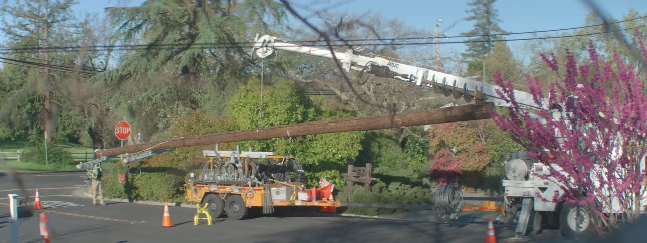 Workers guide a mobile crane that is carrying a tall utility pole