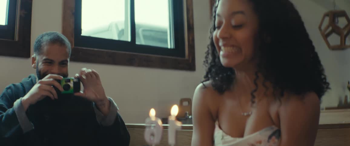 A father is taking a photo of her daughter who is sitting at a table wearing a tiara with a birthday cake that has lit candles