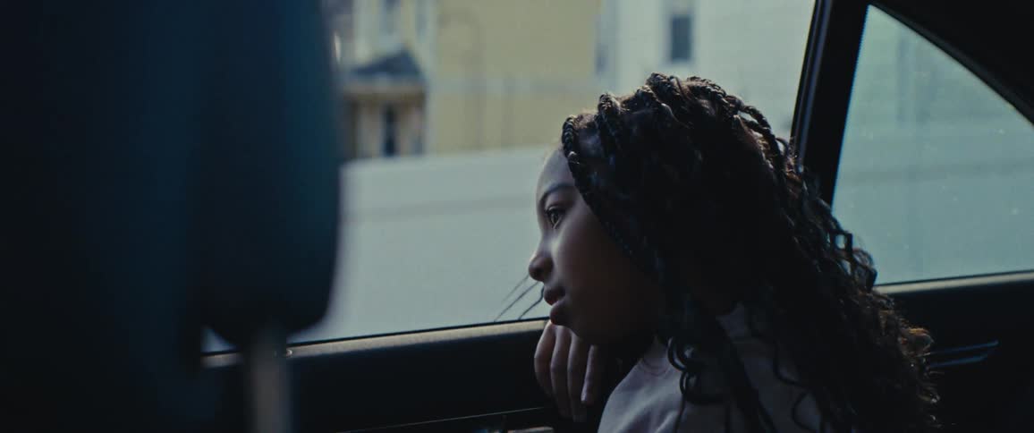 A girl sits in the backseat of a moving car reclined against the window and looking out