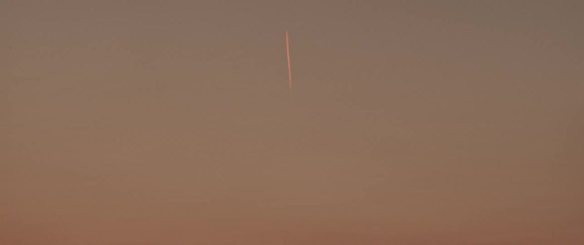 Beginning with a silhouetted landscape field at sunset the camera smoothly transitions upwards revealing and zooming in on an airplane contrail high in the evening sky