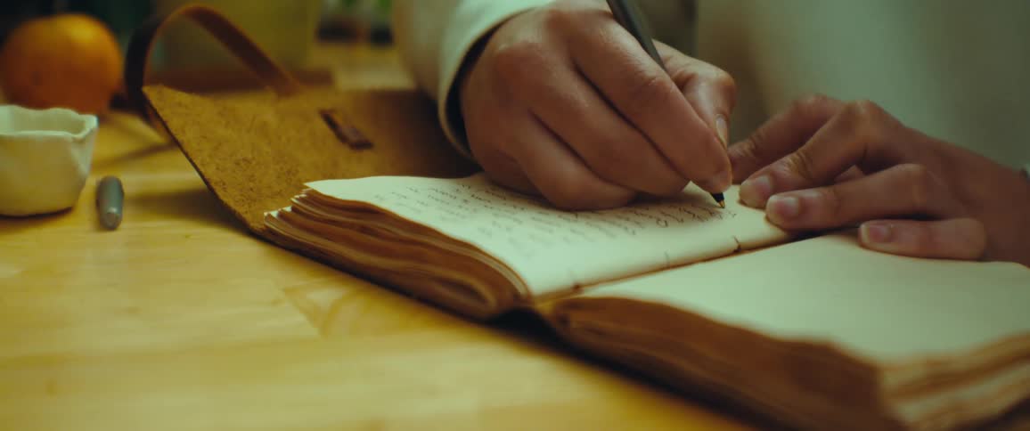 A person writes in an old worn notebook with a pen on a wooden table