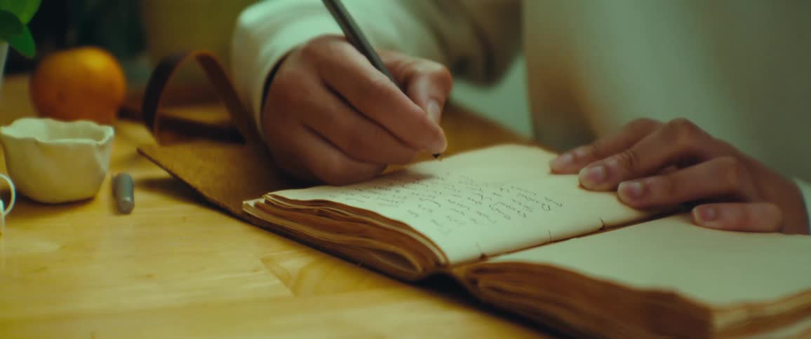 A person writing in a worn open journal with a pencil positioned on a wooden surface beside a small white container and an orange