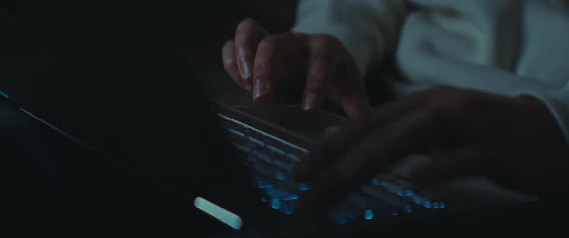 A person wearing a white long sleeved shirt is typing on a laptop keyboard in a dimly lit setting with the glow of the screen reflecting on their hands