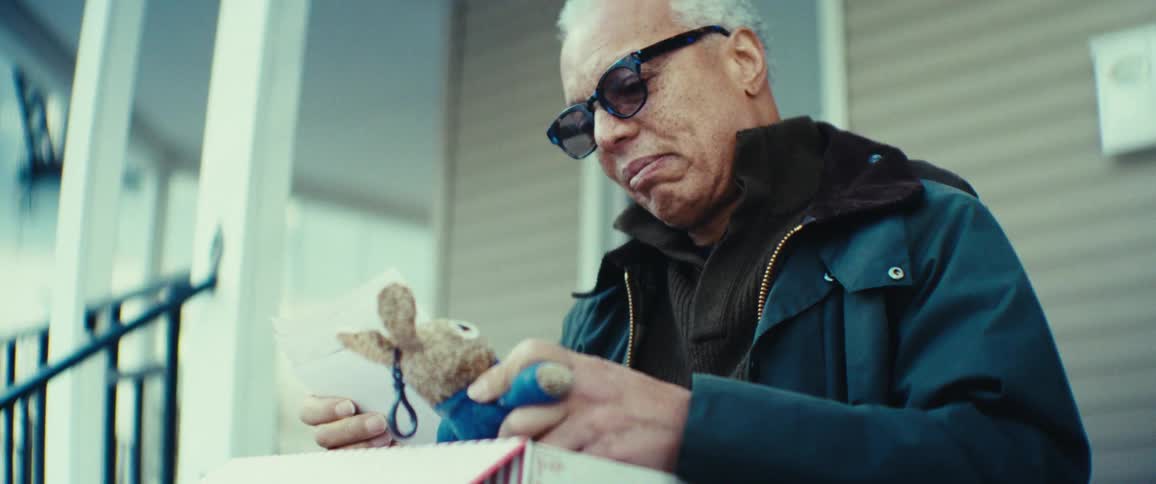 Man wearing sunglasses opens a box and holds a rabbit keychain in front of a house