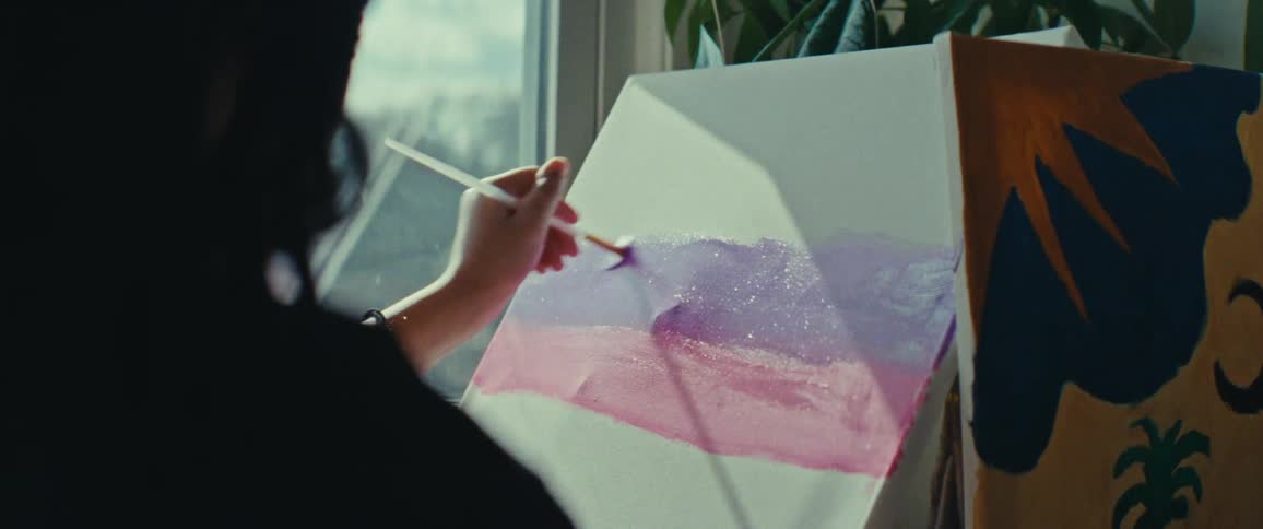 A person is painting on a canvas using a brush creating a gradient of purple and pink colors with a plant visible in the background near a window