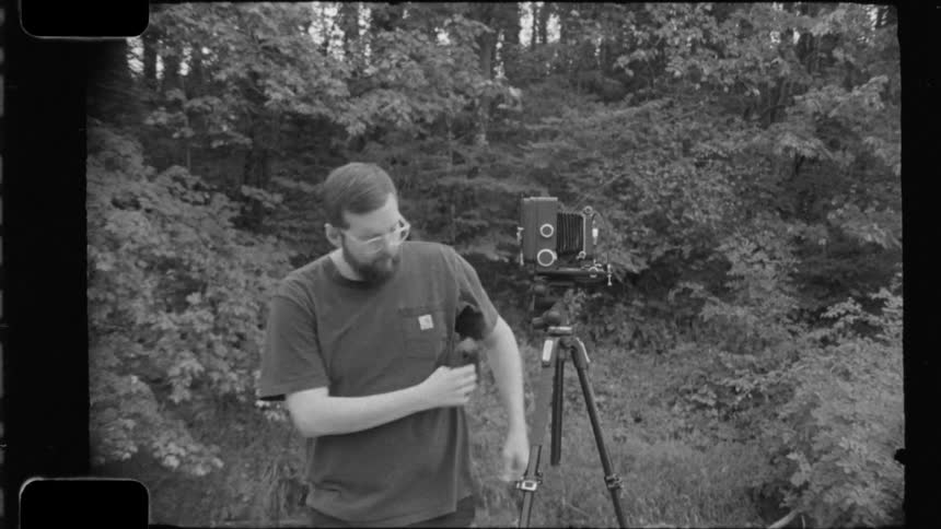 Photographer adjusts his analog camera on a tripod surrounded by a wooded landscape