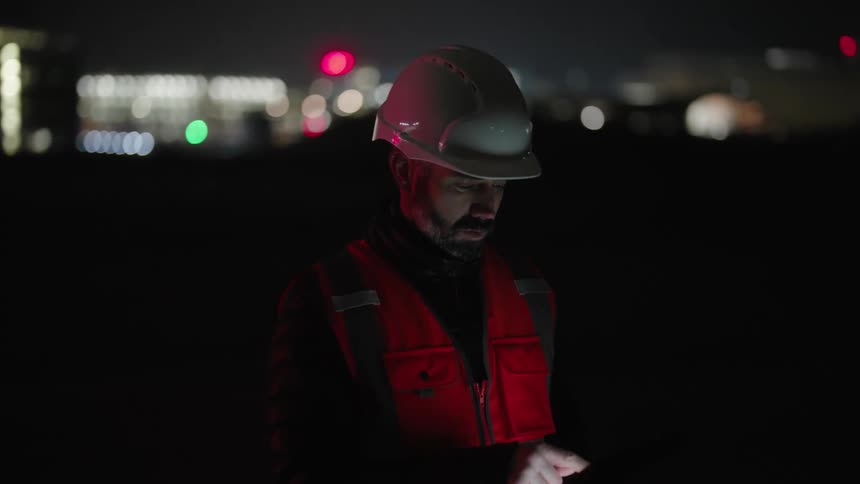 A person wearing a hard hat and a reflective vest uses a tablet at night with an illuminated cityscape in the background