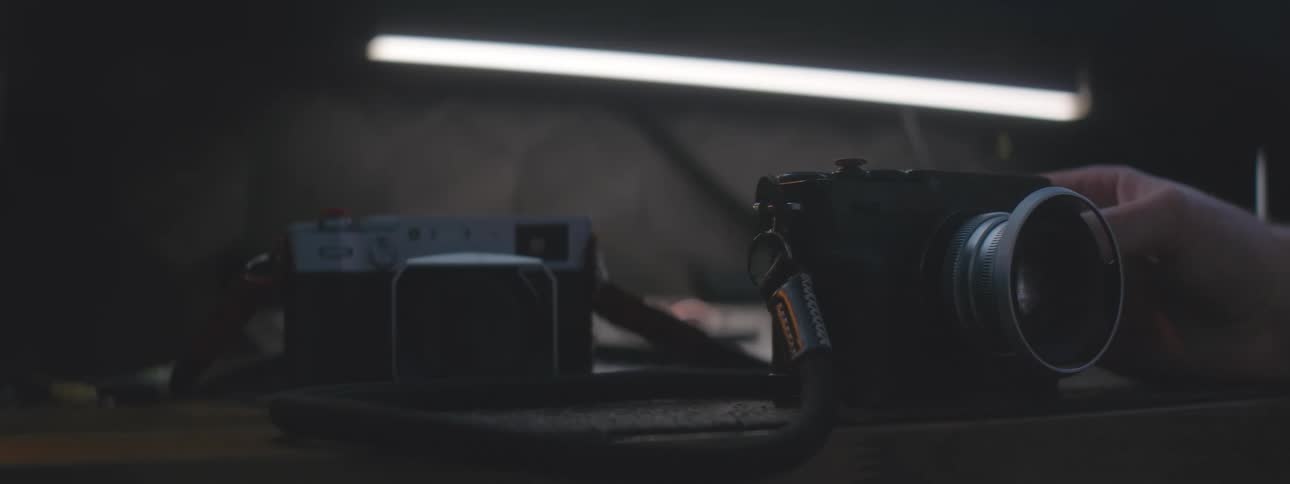 A person carefully handles vintage film cameras over a table in a dimly lit room illuminated by a soft fluorescent light