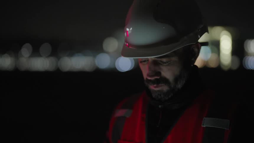 A person wearing a hard hat and a reflective vest uses a tablet at night with an illuminated cityscape in the background