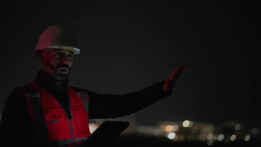 Worker holding a tablet and wearing a helmet on a protective vest while standing in an open air environment at night