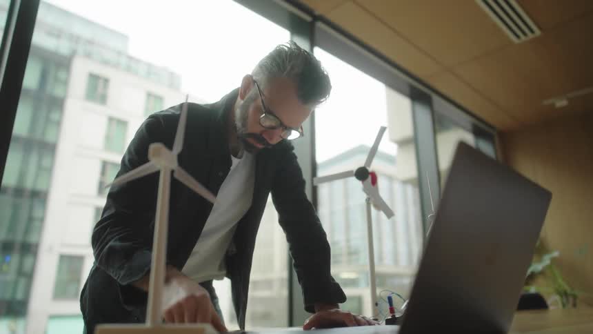 Man putting on glasses and writing in a notebook while working in a modern office with models of wind turbines