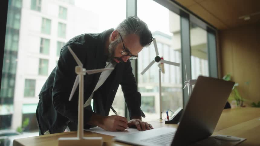A person inspects a small model of a wind turbine while taking notes in an office by a window with a street view