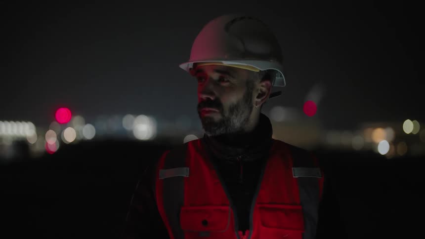 A person wearing a hard hat and a reflective vest uses a tablet at night with an illuminated cityscape in the background