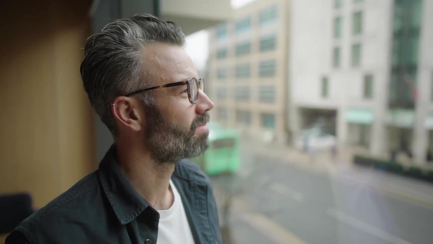 The face of a partially grayhaired man wearing glasses looking through a glass window