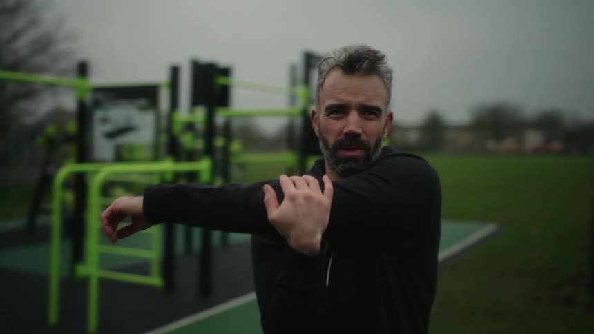 At an outdoor gym a man is doing arm stretches