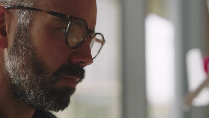 Bearded man with glasses sits at table using a smartphone and a laptop