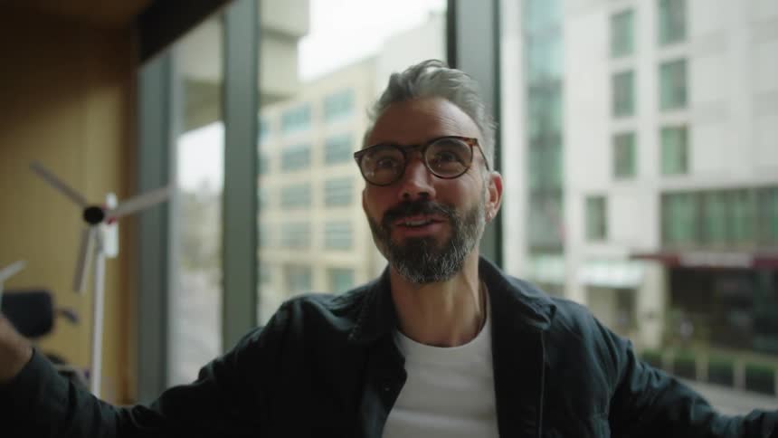 A man sits by a window in an office talking and gesturing during a video call