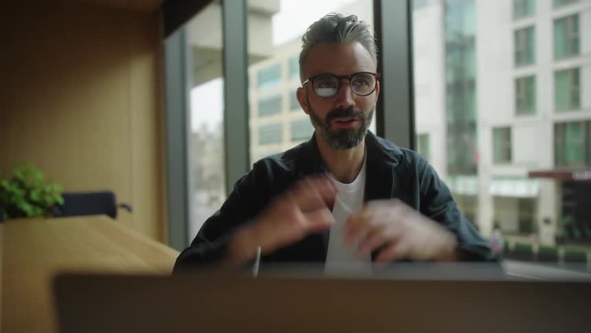 A middle aged man with glasses and a beard is sitting at a desk talking and gesturing enthusiastically during a video call on his laptop with a city street visible outside the window
