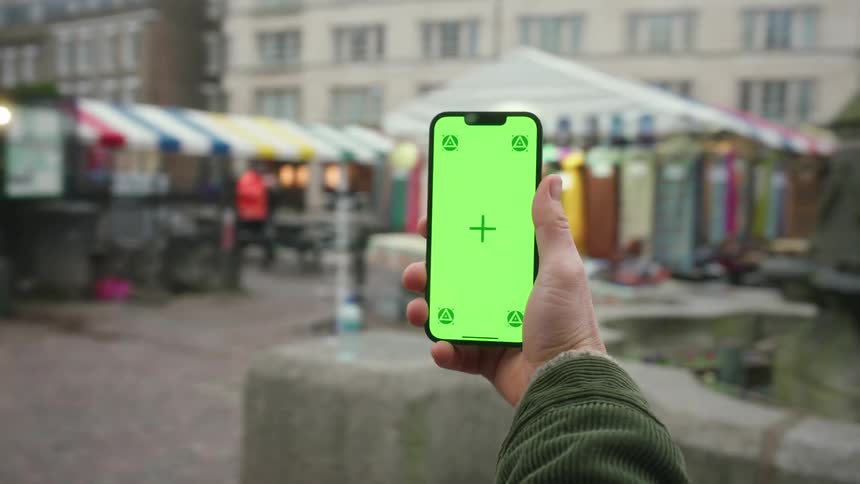 Person holding a smartphone in front of an openair market with stalls and pedestrians