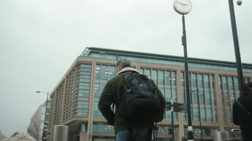 A person in a green jacket with a fur collar carries luggage while walking down the street