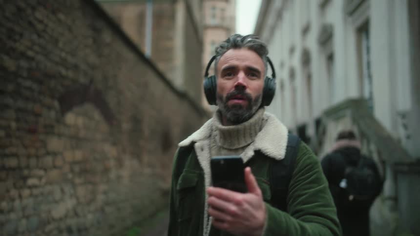 A person wearing headphones and a green jacket with a fur collar is holding a smartphone while walking down a narrow street lined with brick and stone buildings