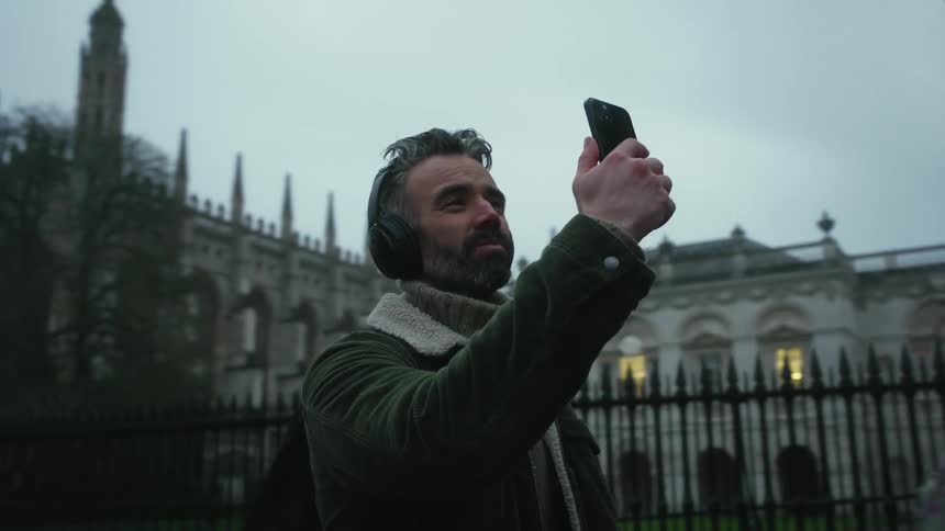 A bearded man in a green jacket with headphones over his head films the city with a smartphone