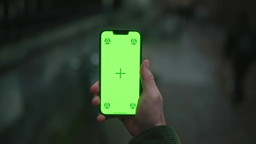 A hand is holding a smartphone with a bright green screen displaying a central plus sign and four triangular symbols in each corner