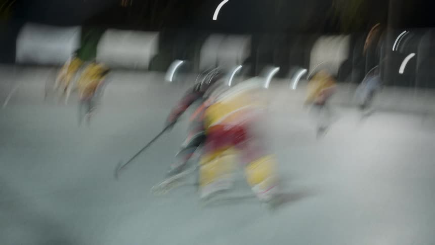 An outdoor nighttime hockey game with players in yellow and red uniforms competing on the rink