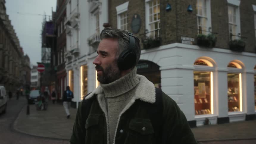 A bearded man in a green jacket with headphones over his head stands gazing at the cityscape
