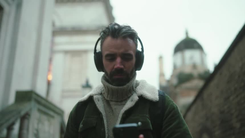 Man holding a smartphone and wearing headphones walking through the city during the day