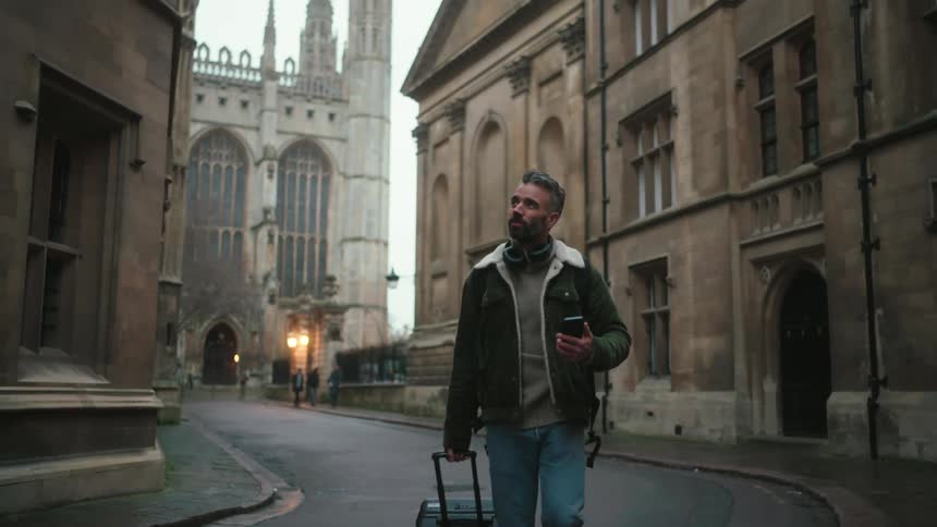 A person wearing headphones and a green jacket with a fur collar is holding a smartphone while walking down a street lined with brick and stone buildings