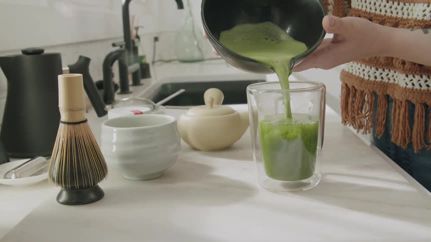 A person pours matcha tea from a dark bowl into a clear glass on a white countertop surrounded by a bamboo whisk a white bowl and a small ceramic pot