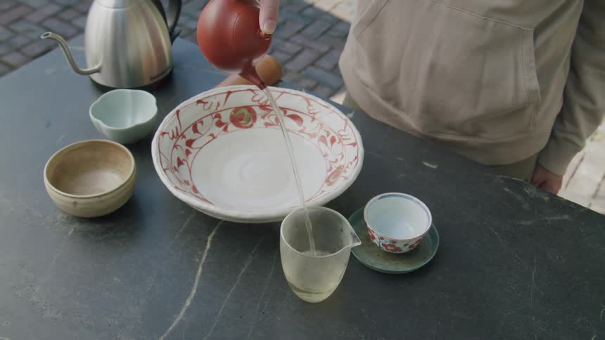 A person is pouring liquid from a red teapot into a glass on a table set with various ceramic bowls and a metal kettle