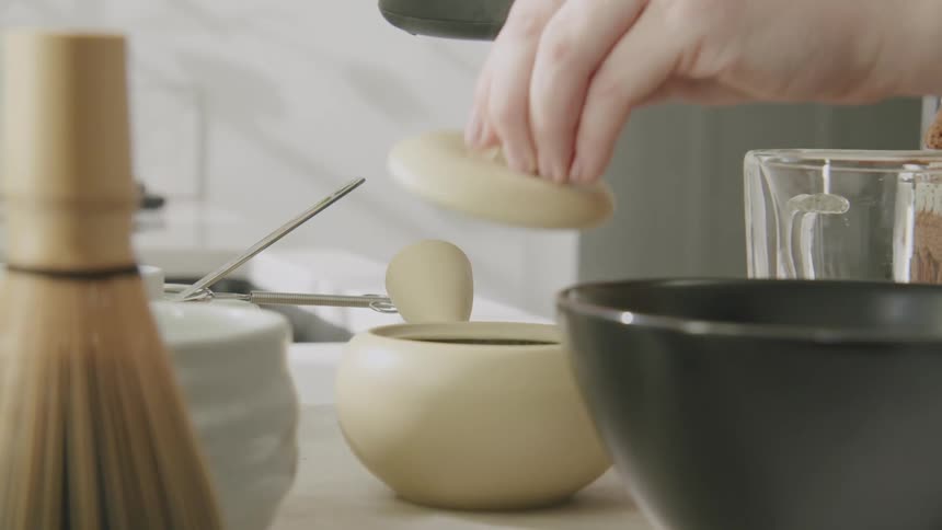 A person pours hot water into a bowl to prepare matcha tea on a white countertop surrounded by a bamboo whisk and ceramic bowls