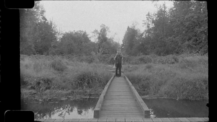 Person with a camera stands at the end of a wooden bridge over a river surrounded by dense vegetation
