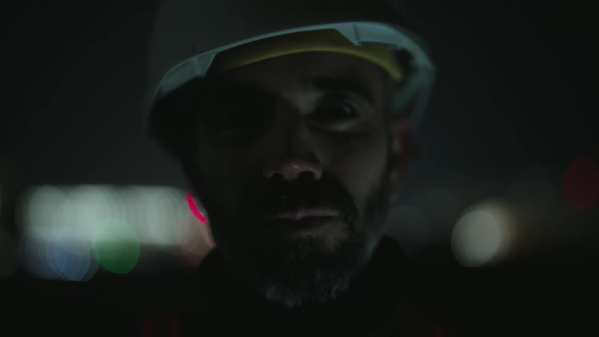 A person wearing a hard hat and a reflective vest looks at the camera with an illuminated cityscape in the background