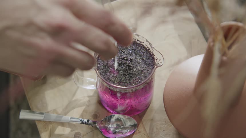 Person adds liquid to a purple mixture in a glass cup on a wooden table