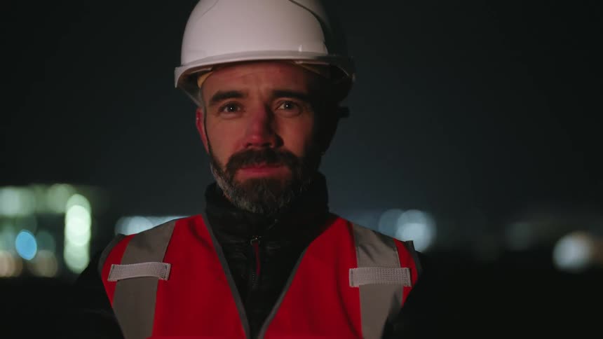 A person wearing a hard hat and a reflective vest at night with an illuminated cityscape in the background