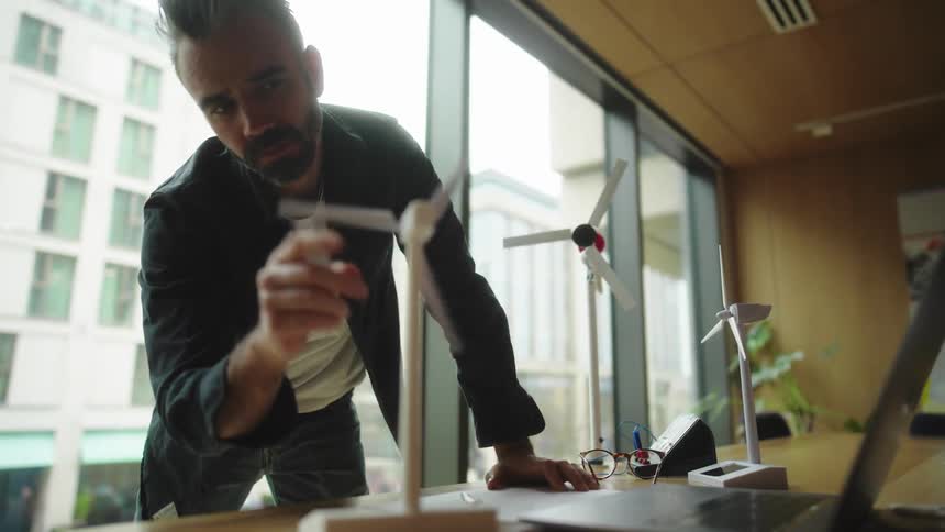 Man adjusts a model wind turbine on a desk in an office with large windows