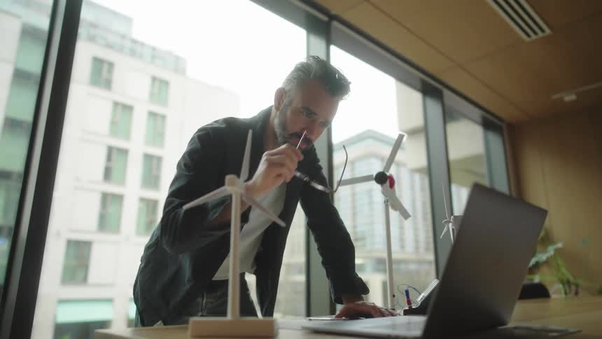 A man standing in front of a laptop puts on reading glasses and starts writing