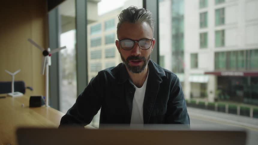 Man gestures and talks during a video conference while sitting in a meeting room with a view of a busy street