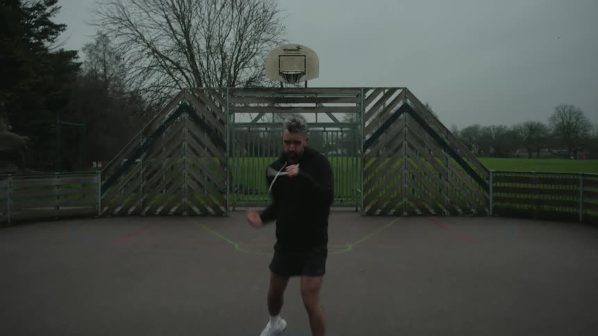 A person boxing on an outdoor basketball court with a metal backboard and hoop surrounded by trees