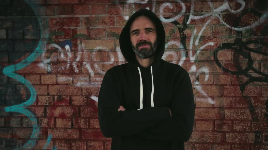 Man in black hooded sweatshirt crosses his arms in front of a graffitied brick wall