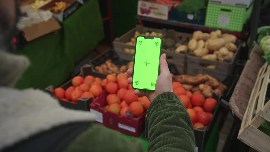 Person holds a smartphone in front of boxes of fruits and vegetables in a market