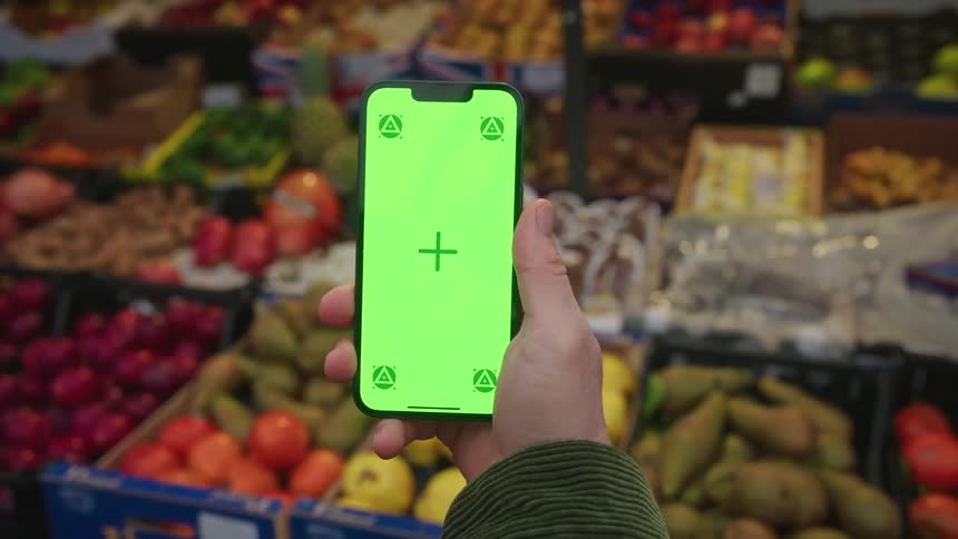 A persons hand holding a cell phone with a green screen and behind there are some boxes of fruit