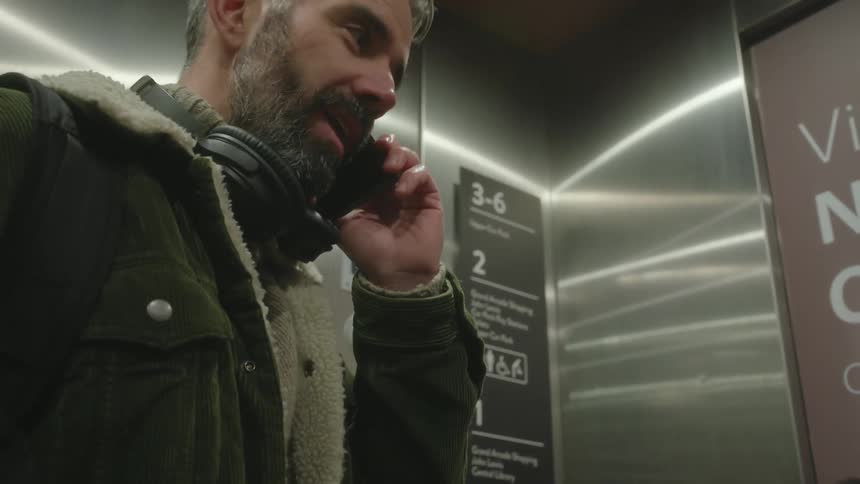 A man in a green jacket with headphones around his neck talks on a smartphone while riding an elevator